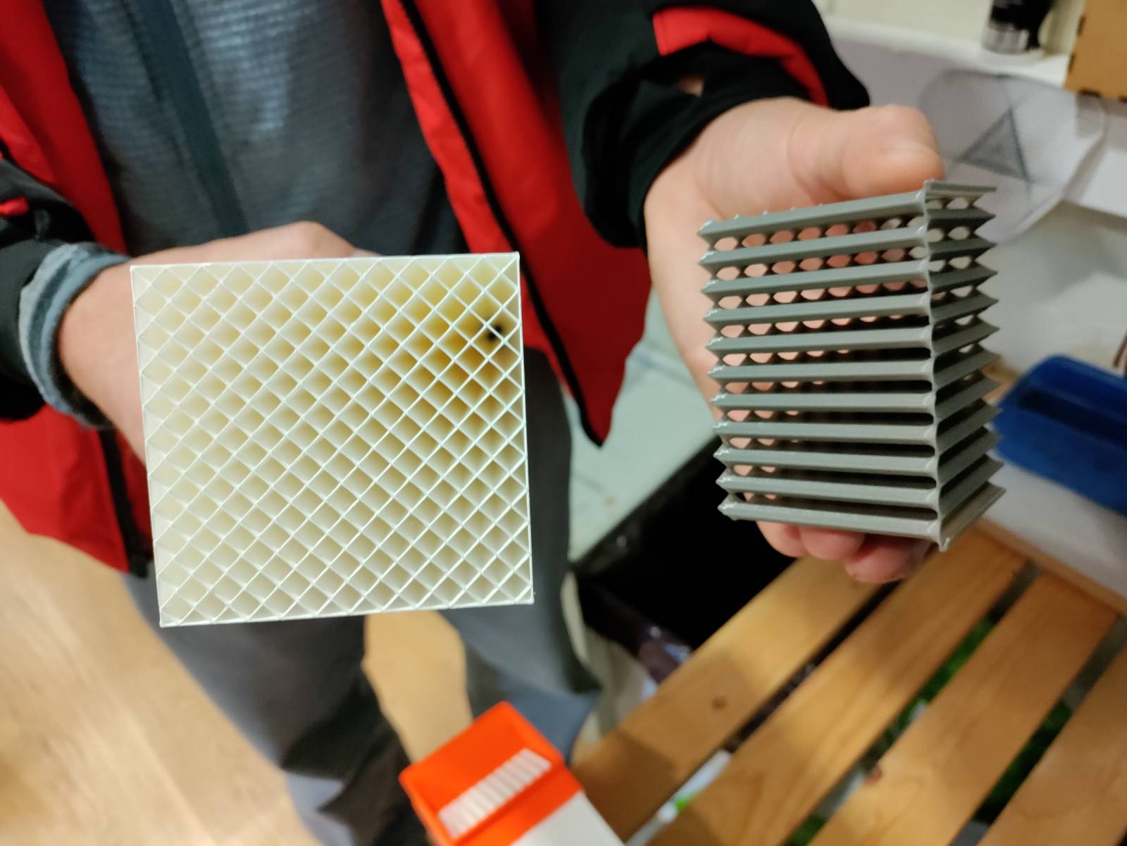 Two hands holding 3D-printed parts: a white square cross-section of the honeycomb tubes, and a grey slotted diverter
